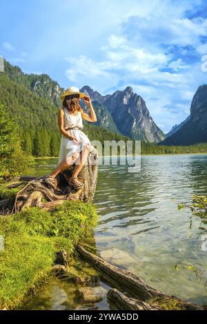 Jeune femme avec chapeau de paille assis sur un tronc d'arbre au bord du lac admirant le paysage de montagne dans les dolomites Banque D'Images