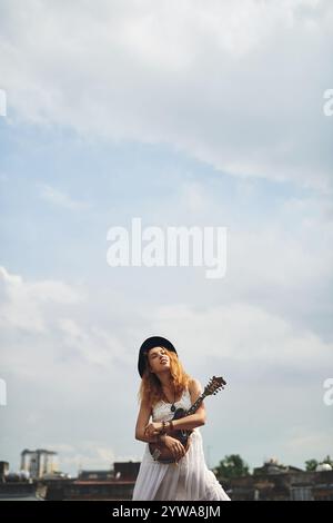 Femme, toit et guitare en ville pour la musique avec réflexion, inspiration et espace maquette avec des nuages dans le ciel. Fille, personne et artiste avec câlin pour Banque D'Images