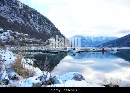 Un port enneigé paisible à Gaupne, en Norvège, avec des bateaux amarrés par une eau calme avec de superbes reflets de montagne en hiver. Banque D'Images