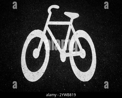 Un grand symbole de vélo blanc est peint sur de l'asphalte foncé, marquant une piste cyclable désignée dans une zone urbaine animée. La lumière du soleil met en valeur le des clair Banque D'Images