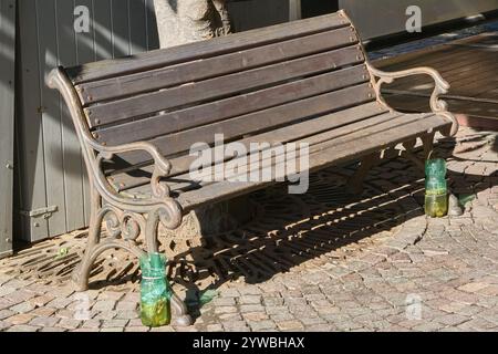 Un banc en bois avec structure en fer forgé situé sur une rue pavée, avec des bouteilles en plastique vert recyclé utilisées comme cendriers à chaque extrémité. Le Banque D'Images