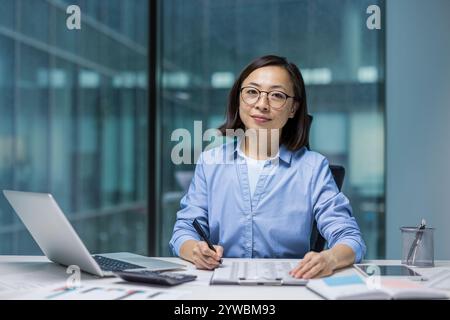 Une femme d’affaires asiatique se concentre sur son travail dans un environnement de bureau contemporain, entourée d’outils nécessaires comme un ordinateur portable et des documents. Elle reflète le professionnalisme et la détermination Banque D'Images
