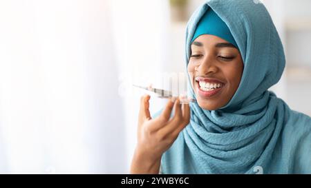 Jeune femme musulmane noire dans le hijab à l'aide de l'assistant vocal virtuel sur smartphone Banque D'Images