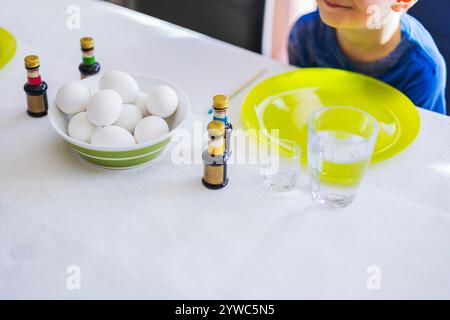 Enfant à table pendant la préparation des oeufs de Pâques, entouré de bouteilles de colorant alimentaire et d'oeufs blancs dans un bol. Suède. Banque D'Images