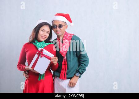 Charmant jeune couple asiatique portant un pull de Noël rouge, chapeau de Père Noël et écharpe, joyeux regardant vers l'avant embrassant portant un cadeau de boîte de Noël blanc, Banque D'Images