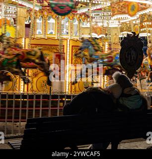 Couple assis sur un banc regardant un carrousel bien éclairé avec des chevaux colorés filant la nuit dans un parc d'attractions Banque D'Images