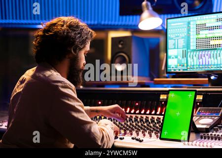 Un technicien audio spécialisé regarde l'écran vert sur l'appareil à côté de la console de mixage dans la salle de contrôle, studio de musique professionnel. Ingénieur du son ajustant les niveaux de volume sur les pistes, post-production. Banque D'Images