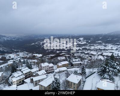 Vue aérienne capturant un village italien enneigé niché parmi les collines ondulantes, soulignant la beauté sereine d'un paysage hivernal sous un nuageux Banque D'Images
