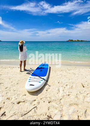 Un voyageur profite de la vue imprenable à Samae San Island Thaïlande, debout près d'un paddleboard sur la côte immaculée. Les eaux bleues claires et le sable doux créent une évasion parfaite de la vie quotidienne. Banque D'Images