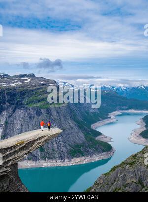 Deux randonneurs se tiennent au bord de la falaise de Trolltunga en Norvège, surplombant un magnifique lac turquoise et un vaste paysage montagneux. Un couple diversifié d'hommes et de femmes en vacances Banque D'Images