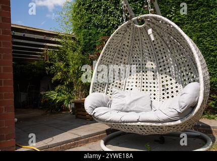 Un fauteuil balançoire en rotin blanc, style oeuf, se tient vide dans le jardin arrière par une chaude journée ensoleillée en Angleterre. Banque D'Images