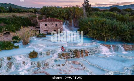 Les visiteurs apprécient les eaux chaudes et riches en minéraux des thermes de Saturnia au coucher du soleil, projetant une lueur dorée sur les cascades et la végétation luxuriante entourant la région. Banque D'Images