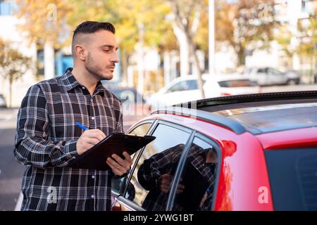 Agent d'assurance écrivant des notes sur le presse-papiers après avoir inspecté une voiture rouge garée dans la rue Banque D'Images