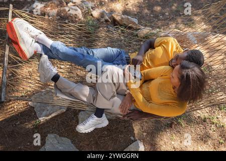 Couple afro-américain s'amusant à regarder des photos de vacances sur un téléphone portable tout en se relaxant dans un hamac entouré par une belle nature. Banque D'Images