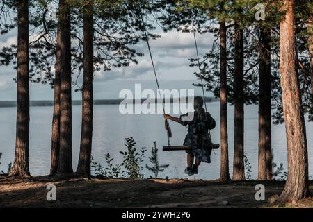 Une femme se balance sur une balançoire d'arbre dans une forêt près d'un lac. La scène est paisible et sereine, avec le bruit de l'eau et le bruissement du Banque D'Images