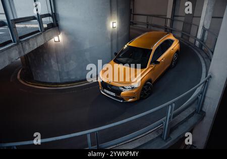 Un VUS orange descend une rampe en spirale dans le parking couvert. Vue de trois quarts avant du crossover français DS7 sur une rampe de sortie de garage. Banque D'Images