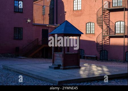Un puits en bois se dresse dans une cour pavée entourée de charmants bâtiments en briques rouges. Le soleil de fin d'après-midi projette des ombres, ajoutant de la chaleur à la Banque D'Images