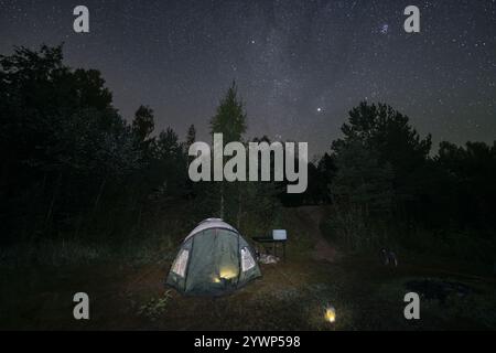 Une tente éclairée de l'intérieur se dresse dans une clairière dans les bois la nuit, le ciel étoilé offrant une toile de fond à couper le souffle. Banque D'Images