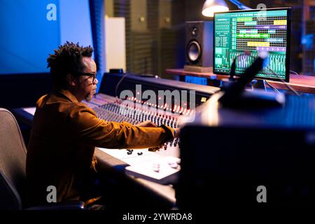 Producteur de disques afro-américain composant des airs dans la salle de contrôle, éditant de la musique et ajustant les niveaux de volume avec les boutons du tableau de bord. Expert audio appuyant sur les curseurs pour ajouter des effets sonores. Banque D'Images