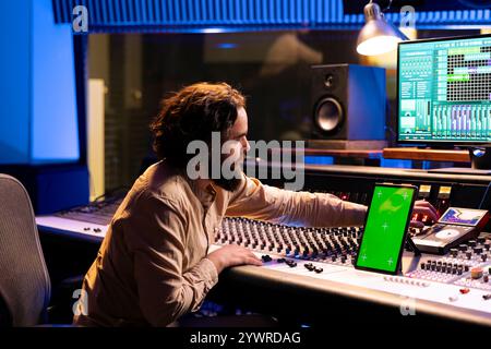 Jeune ingénieur du son appuyant sur les boutons de la console de mixage à côté de l'écran vert sur gadget, enregistrant ses pistes et éditant les niveaux de volume. Panneau de commande de technicien de producteur de musique. Banque D'Images