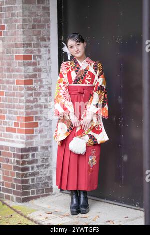 Une femme japonaise dans la vingtaine, portant un kimono rouge (hakama), un incontournable des diplômés universitaires japonais, se tient devant la grande porte de fer noir de Banque D'Images