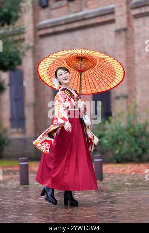 Une japonaise d'une vingtaine d'années, portant un kimono rouge (hakama), un incontournable des diplômés d'université japonais, sourit et pose sous la pluie avec un himono japonais Banque D'Images