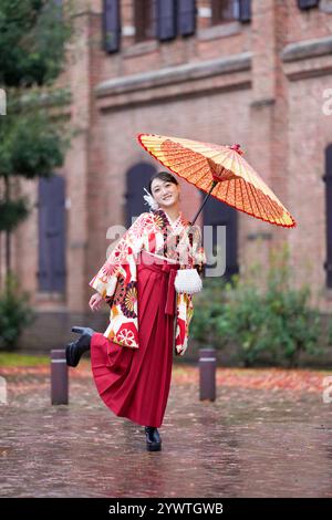 Une japonaise d'une vingtaine d'années, portant un kimono rouge (hakama), un incontournable des diplômés d'université japonais, sourit et pose sous la pluie avec un himono japonais Banque D'Images