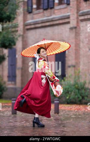 Une japonaise d'une vingtaine d'années, portant un kimono rouge (hakama), un incontournable des diplômés d'université japonais, sourit et pose sous la pluie avec un himono japonais Banque D'Images