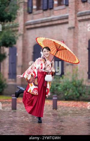 Une japonaise d'une vingtaine d'années, portant un kimono rouge (hakama), un incontournable des diplômés d'université japonais, sourit et pose sous la pluie avec un himono japonais Banque D'Images