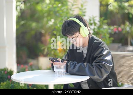 Une femme chinoise dans les années 30 en vêtements sportifs utilise un smartphone tout en écoutant de la musique à travers des écouteurs à une table dans un café élégant sur Jilu Ro Banque D'Images