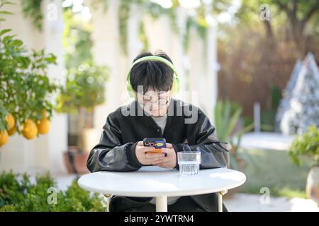 Une femme chinoise dans les années 30 en vêtements sportifs utilise un smartphone tout en écoutant de la musique à travers des écouteurs à une table dans un café élégant sur Jilu Ro Banque D'Images