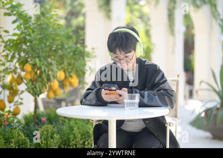 Une femme chinoise dans les années 30 en vêtements sportifs utilise un smartphone tout en écoutant de la musique à travers des écouteurs à une table dans un café élégant sur Jilu Ro Banque D'Images