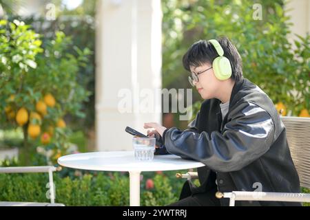 Une femme chinoise dans les années 30 en vêtements sportifs utilise un smartphone tout en écoutant de la musique à travers des écouteurs à une table dans un café élégant sur Jilu Ro Banque D'Images