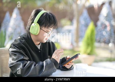 Une femme chinoise dans les années 30 en vêtements sportifs utilise un smartphone tout en écoutant de la musique à travers des écouteurs à une table dans un café élégant sur Jilu Ro Banque D'Images