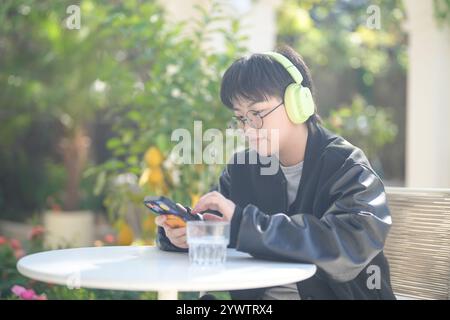 Une femme chinoise dans les années 30 en vêtements sportifs utilise un smartphone tout en écoutant de la musique à travers des écouteurs à une table dans un café élégant sur Jilu Ro Banque D'Images