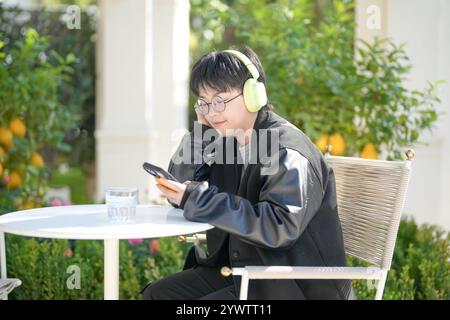 Une femme chinoise dans les années 30 en vêtements sportifs utilise un smartphone tout en écoutant de la musique à travers des écouteurs à une table dans un café élégant sur Jilu Ro Banque D'Images