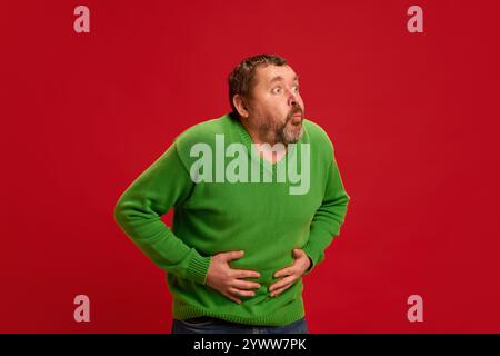 Photo d'un homme mature habillé de façon désinvolte tenant les mains sur le ventre et la bouche ouverte, ressentant des maux d'estomac et des nausées sur fond de studio rouge. Banque D'Images