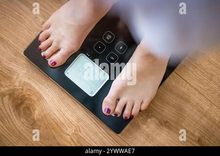 Vue rapprochée d'une femme pieds nus sans visage debout sur une balance numérique avec un affichage pour mesurer le poids et la graisse corporelle sur un plancher en bois de chêne Banque D'Images