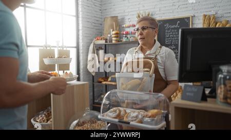 Femme travaillant dans la boulangerie parlant à l'homme client dans une pâtisserie intérieure remplie de pain frais et de pâtisseries colorées Banque D'Images