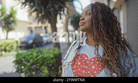 Jeune femme afro-américaine avec des cheveux tressés portant une chemise à motif de coeur, debout dans une rue urbaine, regardant réfléchie et confiante dans un cit Banque D'Images