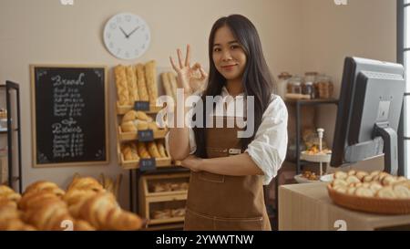 Jeune femme chinoise dans une boulangerie donne un geste de la main OK tout en se tenant près du comptoir avec du pain frais et des pâtisseries dans un intérieur de magasin confortable Banque D'Images