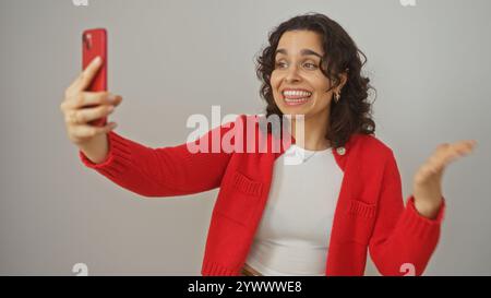 Une jeune femme hispanique dans un pull rouge s'engageant joyeusement dans un appel vidéo sur un fond blanc isolé. Banque D'Images