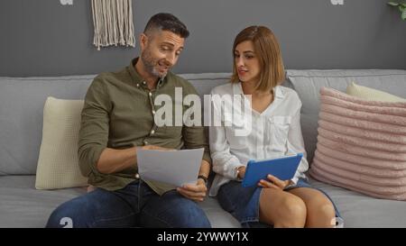Homme et femme assis sur un canapé dans le salon, examinant des documents et utilisant une tablette dans un cadre confortable à la maison. Banque D'Images