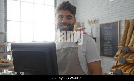 Jeune homme hispanique avec une barbe souriant derrière le comptoir d'une boulangerie intérieure remplie de pain et de pâtisseries Banque D'Images