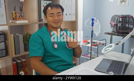 Jeune vétérinaire chinois portant des gommages verts, se pointant vers lui-même et souriant dans un bureau de clinique vétérinaire avec un ordinateur portable et un équipement médical Banque D'Images