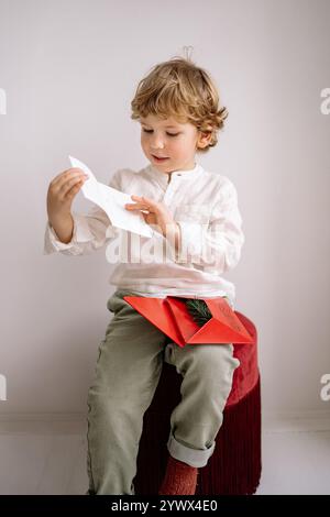 Enfant ouvre avec impatience une lettre de Noël assis sur un tabouret confortable pendant la saison des fêtes Banque D'Images