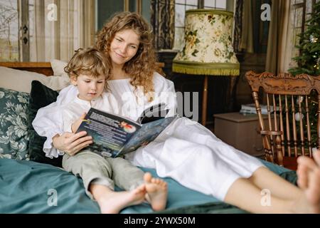 Mère et enfant profitant d'un moment de Noël confortable lisant un livre ensemble dans un salon joliment décoré Banque D'Images