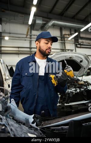 Un jeune mécanicien concentré tient des outils électriques tout en inspectant une voiture dans un environnement d'atelier bien éclairé Banque D'Images