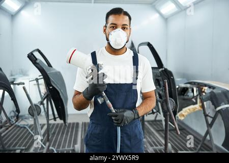 Beau mécanicien portant un masque et des gants se prépare à peindre des pièces de voiture dans un atelier. Banque D'Images