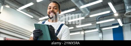 Beau mécanicien sourit tout en utilisant une tablette, mettant en valeur l'expertise dans un atelier de réparation automobile moderne. Banque D'Images